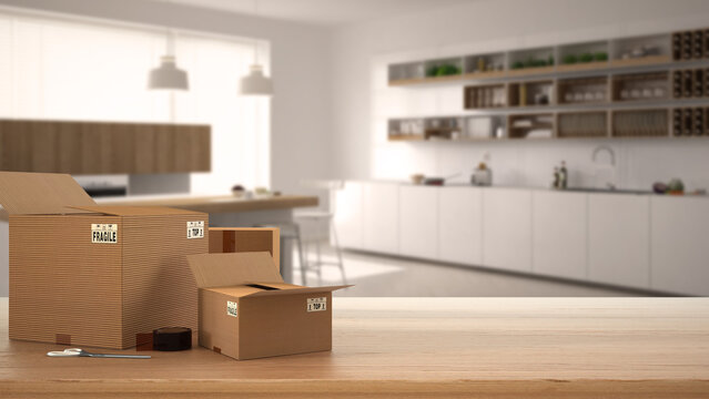 Wooden table, desk or shelf with stack of cardboard boxes over blurred view of minimalist white kitchen with island, modern interior design, moving house concept with copy space