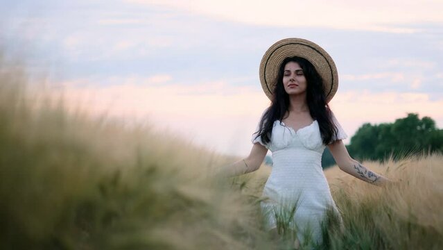 Stunning brunette woman in hat walking down in the wheat field enjoying freedom. Young beautiful brunette woman in a white dress in field. 