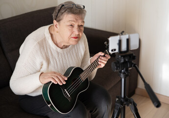 Elderly woman 70+ sits on the couch and learns to play ukulele at home using video tutorial on her smartphone. Video course on learning to play the guitar. Adaptation of pensioners in the modern world