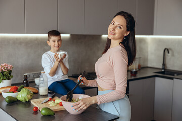 Portrait of beautiful Caucasian young mother cooking salat in kitchen. Small son on background.