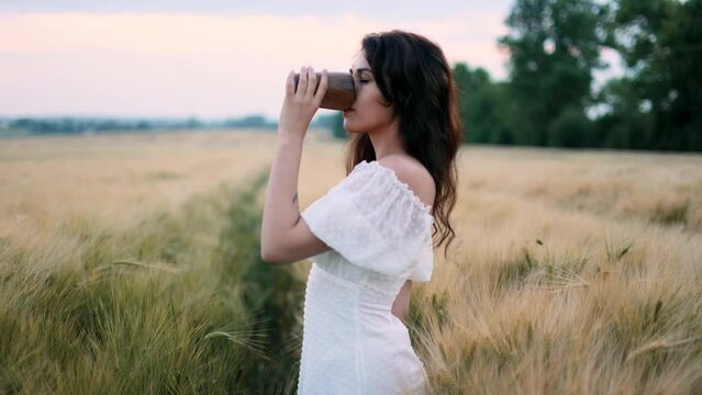 Young woman enjoying coffee or tea from eco wooden cup on warm summer evening sunset in wheat field, concept of harmony woman and nature, healthy lifestyle.