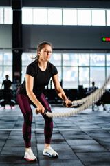 Fototapeta na wymiar Powerful woman training battle ropes at cardio workout in dark gym. Professional athlete exercise fitness sport club equipment. Strong bodybuilder lifting weights. Athletic person effort