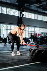 Powerful man using hammer for strength workout in fitness gym. Muscular man hitting big tire with sledgehammer. Strong sportsman practicing intense training in sport club. Silhouette athlete exercise