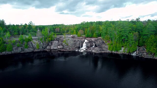 Gushing waterfall flows down rock escarpment into dark colored lake with lush green forest view from above in Northern Ontario