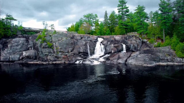 Rushing waterfall cascades through natural bedrock cliff in Northern Ontario with forest and cloudy sky