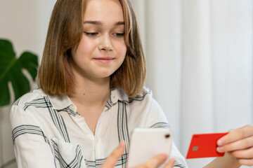 Female professional doing online payment through credit card while sitting at home office. Cheerful young girl shopping or banking online using mobile phone and credit card, living room interior.