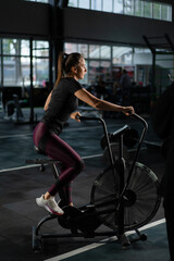 Obraz na płótnie Canvas Young Athletic middle aged woman training on air resistance bike, cross training workout set in gym. Active woman spinning a air bike in gym with trainers. female training on air bike.