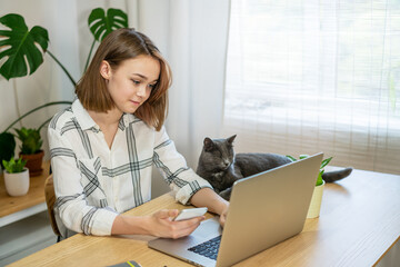 Young woman entrepreneur working on laptop at home with her cat, sitting together in modern room with plants. Girl using a computer for study online at home, female user busy on a distance internet