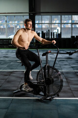 Naklejka premium Young Athletic middle aged man training on air resistance bike, cross training workout set in gym. Active man spinning a air bike in gym with trainers. Male training on air bike.