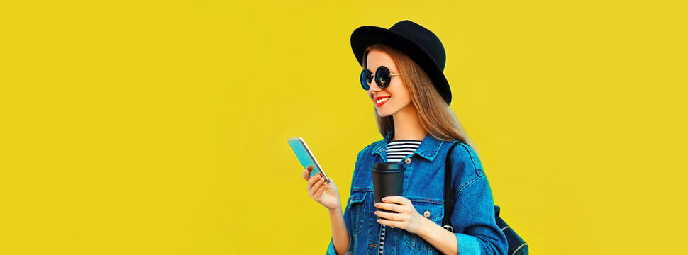 Portrait of stylish happy smiling young woman model with smartphone wearing black round hat, jean jacket on yellow background, blank copy space for advertising text