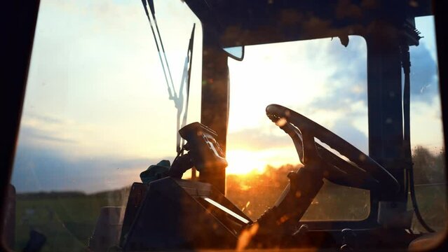 Side view through window of tractor driver cab with steering. Car is standing on field against sunset. Management agriculture with help of equipment. Simplification of production process on farm.