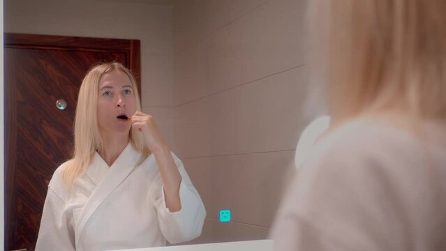 Beautiful young woman in white coat or pajamas brushes her teeth in front of mirror in bathroom. Mirror image of beautiful woman washing her teeth is daily concept of oral hygiene. Fresh good morning