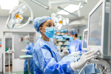 Anesthetist Working In Operating Theatre Wearing Protecive Gear checking monitors while sedating patient before surgical procedure in hospital - Powered by Adobe