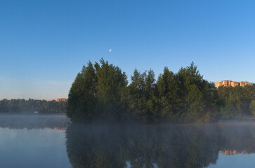 Bright blue sky. Trees near water. Tree reflection on water surface. Transparent mist. Nature background.