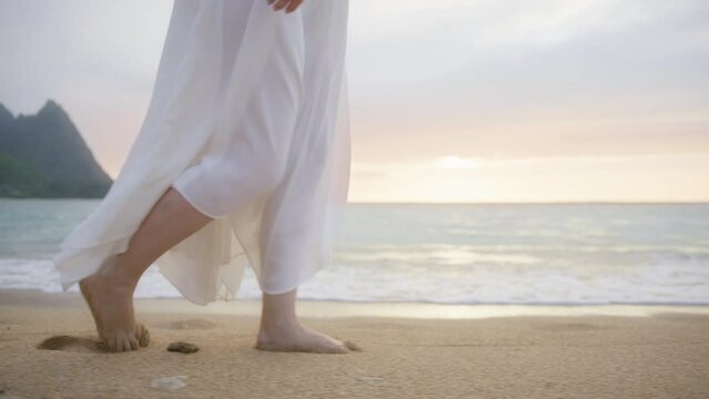 Slow motion woman in white boho dress walking barefoot by beach at gentle golden sunset above horizon leaving footprints in sand. Female tourist on summer vacation in Kauaii, Hawaii island USA travel