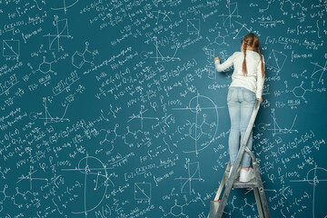 Portrait of happy kid girl resolving complex math problem on blue chalkboard. Kids math education...