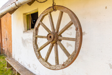 Old wooden carriage wheel with metal fittings hangs on the white wall. Outdoor natural day light.