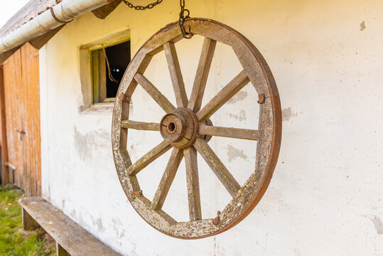 Old wooden carriage wheel with metal fittings hangs on the white wall. Outdoor natural day light.