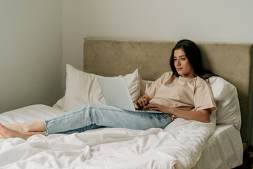 Young brunette freelancer in t-shirt and jeans lying on bed working on laptop