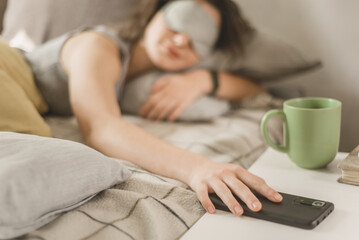 Obraz na płótnie Canvas A girl in a sleep mask and pajamas reaches out to her smartphone to turn off the alarm. morning awakening
