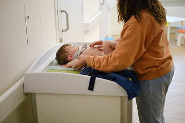 A mother woman puts clothes on a toddler baby on a changing table in the corridor of the clinic....