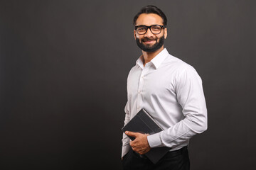Indian male entrepreneur in formal white shirt and glasses carrying laptop isolated on black,...