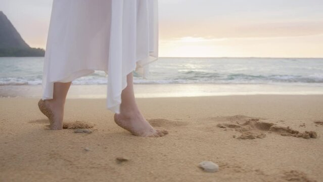 Barefoot female tourist walking tiptoe. Summer vacation on Hawaii island travel. Slow motion woman in white dress walking on tiptoe by beach at golden sunset above horizon leaving footprints in sand