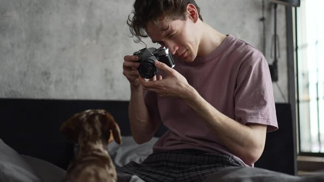 Young man is taking photo of cute pet and sitting on bed in home bedroom spbd. 4k Close view of caucasian guy holds camera in his hand and takes picture of purebred dog, looks with smile and sits in