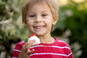 Cute child, boy, eating small cupcake of Pavlova desert, light egg and sugar desert with cream and strawberries