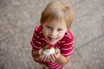 Cute child, boy, eating small cupcake of Pavlova desert, light egg and sugar desert with cream and strawberries