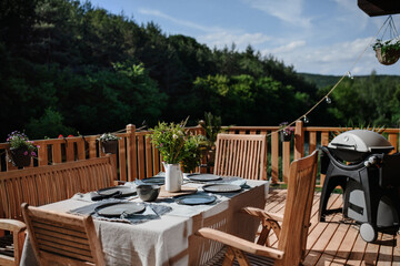 Dining table with wooden chairs set for dinner on the terrace with grill in summer, garden party. concept.