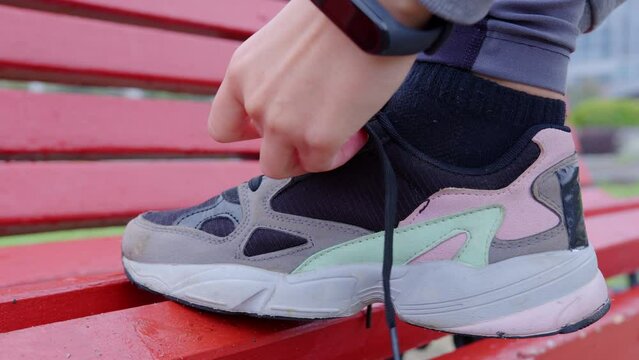 Female hands tying shoelace on sneaker placing her foot on red bench. Faceless girl preparing for jogging or training outdoors. Unrecognizable young woman going to running in sports shoes at street