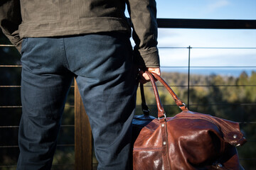 caucasian man behind holding brown leather travel overnight bag blue pants green jacket outdoors view mountains