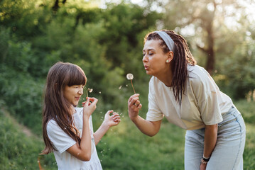 Mother and daughter blowing dandelion during an outdoor walk, smiling and having fun