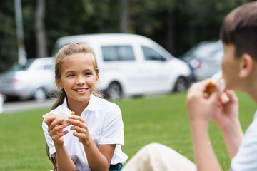 Smiling schoolgirl holding sandwich near blurred classmate in park.