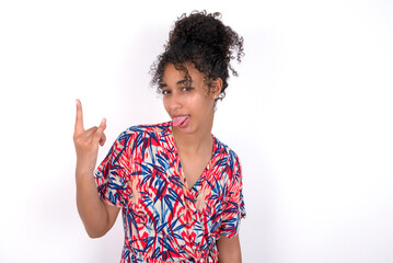 Portrait of a crazy Young African American woman wearing colourful dress over white wall showing tongue horns up gesture, expressing excitement of being on concert of band.