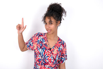 Pleasant looking Young African American woman wearing colourful dress over white wall has clever expression, raises one finger, remembers himself not to forget tell important thing.