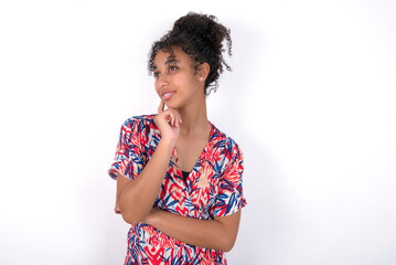 Face expressions and emotions. Thoughtful Young African American woman wearing colourful dress over white wall holding hand under his head, having doubtful look.
