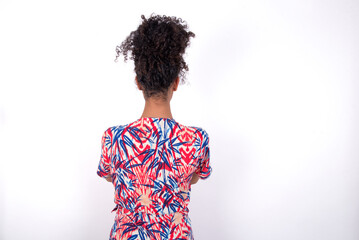Young African American woman wearing colourful dress over white wall standing backwards looking away with arms on body.