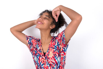 Young African American woman wearing colourful dress over white wall relaxing and stretching, arms and hands behind head and neck smiling happy