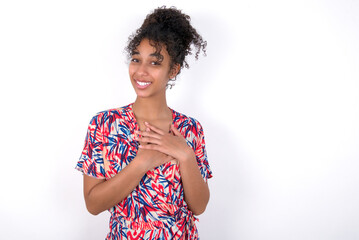 Happy smiling Young African American woman wearing colourful dress over white wall has hands on chest near heart. Human emotions, real feelings and facial expression concept.