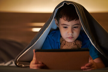 Pensive boy lying under blanket late at night and watching animated cartoon on digital tablet...