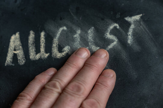 The semi-erased word AUGUST on the black chalkboard. An adult man's left hand removes the handwritten word with his fingers. Fuzzy letters on black surface. End of calendar month concept - Powered by Adobe