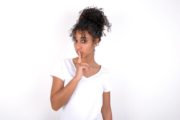 Fototapeta na wymiar Surprised Young beautiful girl with afro hairstyle wearing white t-shirt over white wall makes silence gesture, keeps finger over lips and looks mysterious at camera