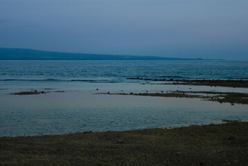 Foggy blue remote shore with pink haze, dark shore and calm sea on sunset, low tide. Tropical indonesian landscape with islands, tourism in asia.