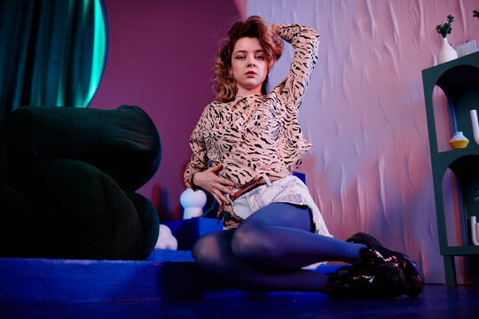 Full length portrait of sensual young woman sitting on floor in club and posing vogue style in neon light, copy space