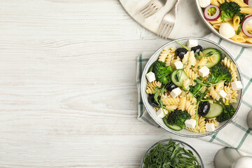 Bowl of delicious pasta with cucumber, olives, broccoli and cheese on white wooden table, flat lay....