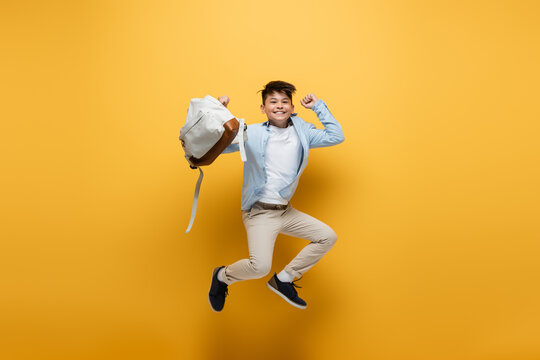 Cheerful asian schoolkid holding backpack and jumping on yellow background.