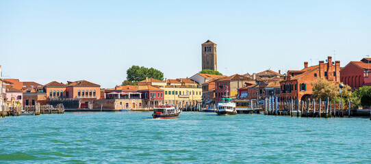 Cityscape of Murano island, famous for the production of artistic glass. Sea channel with two ferry boats, bell tower of the Cathedral, Basilica of saints Maria and Donato, VII century. Venice, Italy. - obrazy, fototapety, plakaty