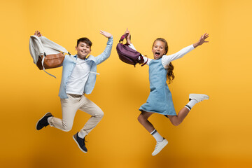 Fototapeta na wymiar Excited multiethnic kids with backpacks jumping on yellow background.